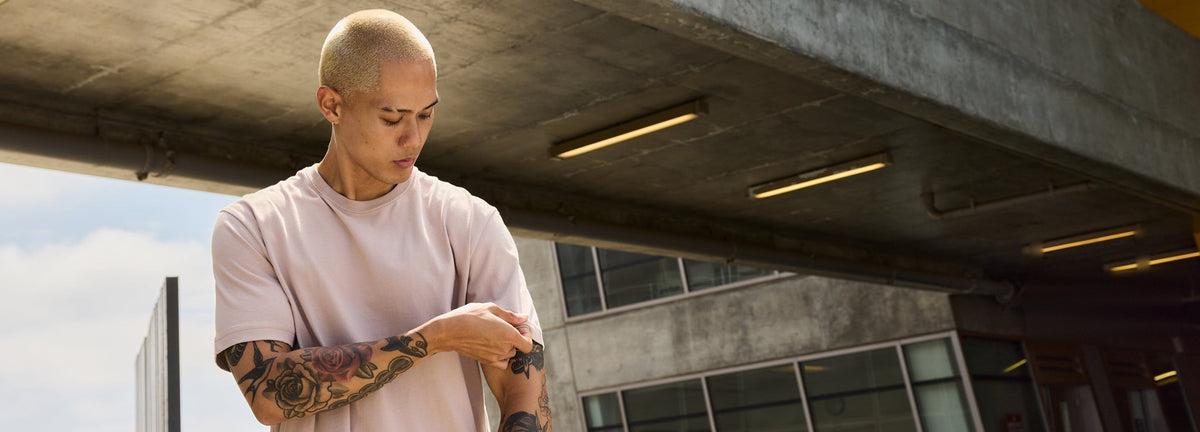 Man in a pale pink Essential Tee rolling up his sleeve under a concrete overpass